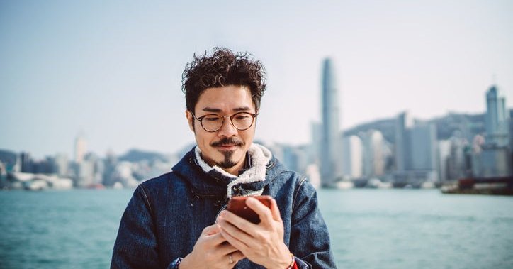 A man looking at his phone while standing along a waterfront with a city skyline behind him.