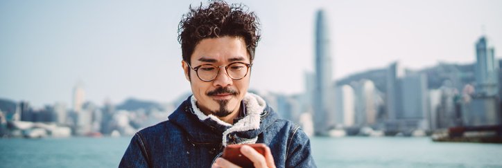 A man looking at his phone while standing along a waterfront with a city skyline behind him.