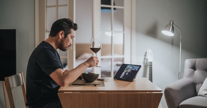 A man drinking wine and eating dinner at home while on a video call with friends.