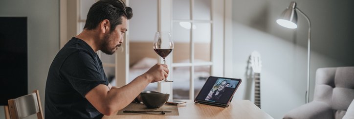 A man drinking wine and eating dinner at home while on a video call with friends.
