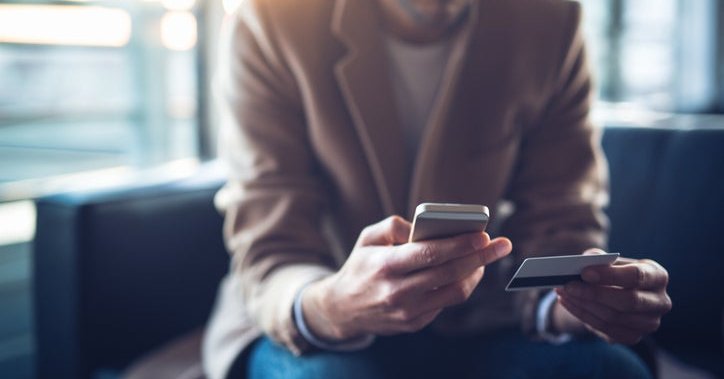 A man sitting on a couch and holding a credit card in one hand and a phone in the other.