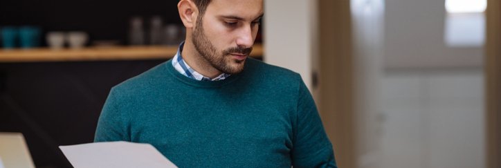 A man standing in his living room and looking between two pieces of paper held in each hand.