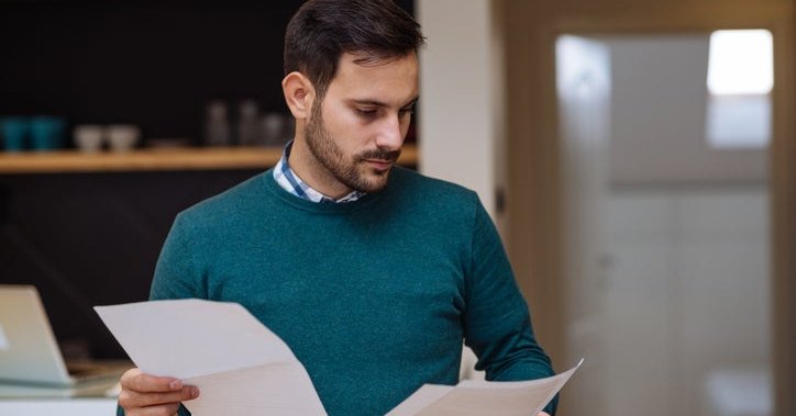 A man standing in his living room and looking between two pieces of paper held in each hand.