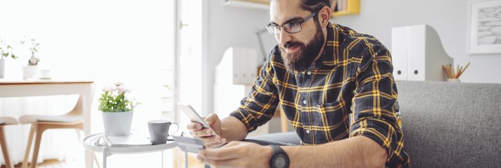 A man sitting on his couch and holding a credit card while looking up something on his phone.