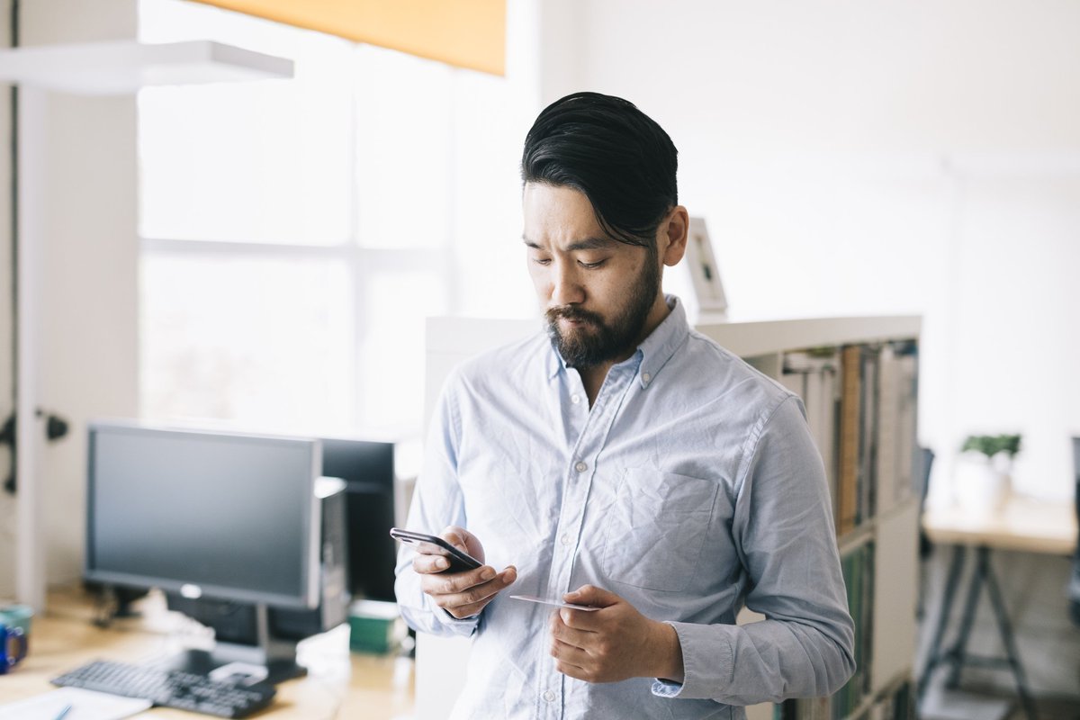 A man standing in an office while holding his phone and a credit card.