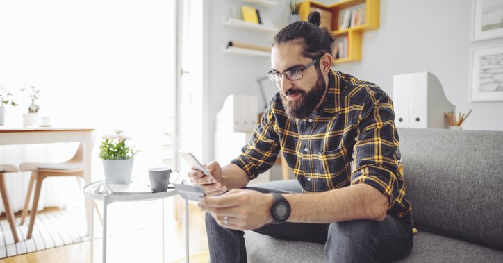 A man sitting on his couch and holding a credit card while looking up something on his phone.