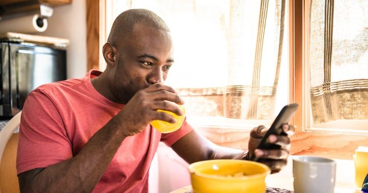 A smiling man drinking juice and looking at his phone while sitting at the breakfast table in front of a bowl of cereal.