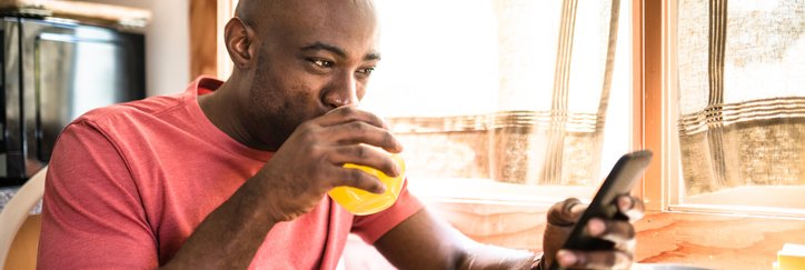 A smiling man drinking juice and looking at his phone while sitting at the breakfast table in front of a bowl of cereal.
