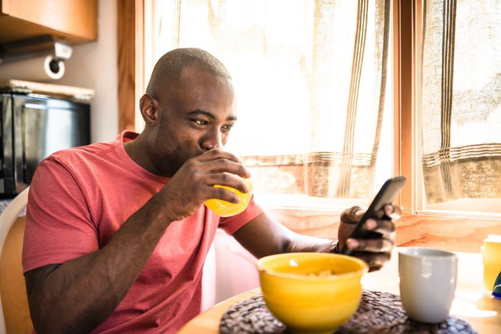 A smiling man drinking juice and looking at his phone while sitting at the breakfast table in front of a bowl of cereal.