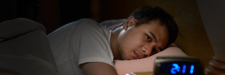 A man lying awake in bed in the dark looking at a clock that reads 2:11.