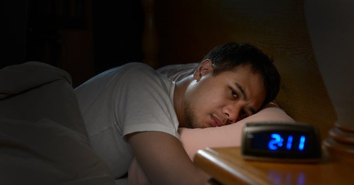 A man lying awake in bed in the dark looking at a clock that reads 2:11.