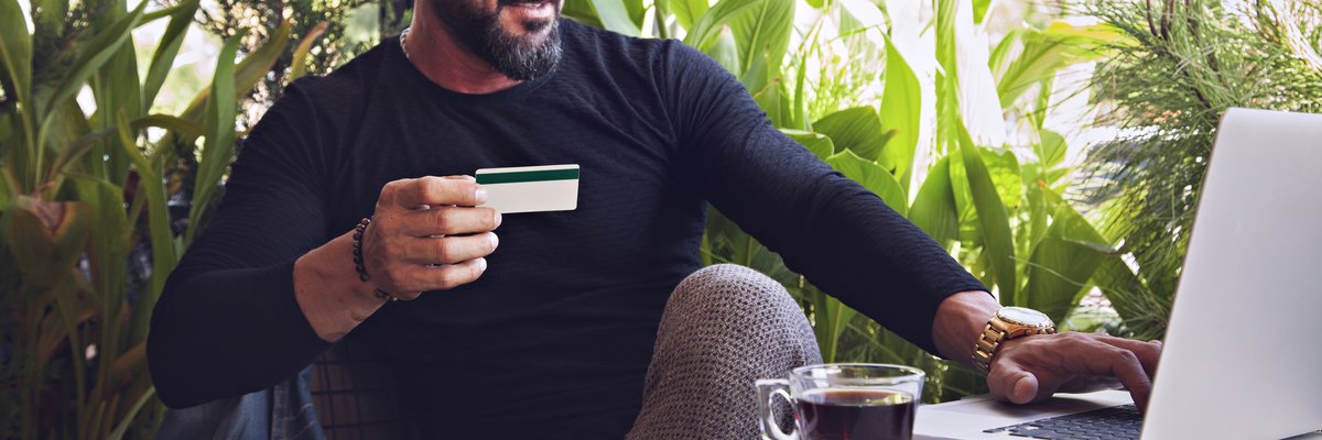 A man sitting in a coffee shop in front of lots of plants working on his laptop and holding a credit card.
