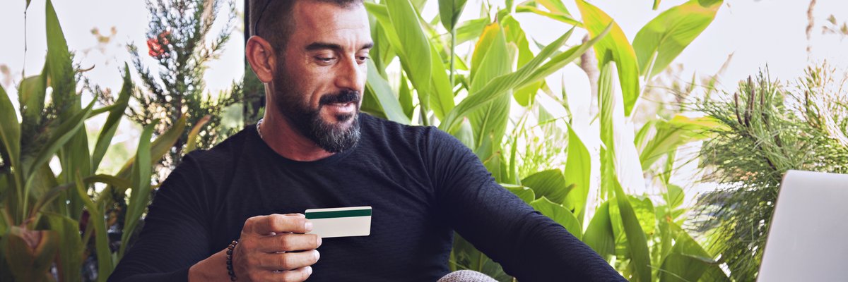 A man sitting in a coffee shop in front of lots of plants working on his laptop and holding a credit card.