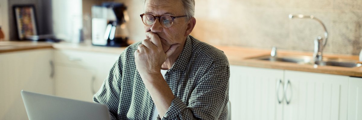 A man sitting in his kitchen and searching on a laptop while resting his chin in his hand.