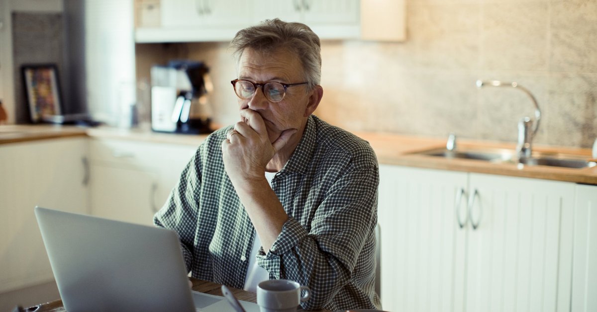 A man sitting in his kitchen and searching on a laptop while resting his chin in his hand.