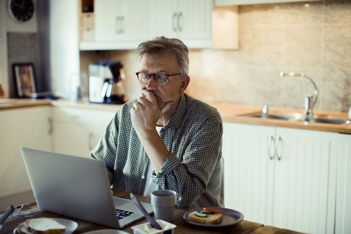 A man sitting in his kitchen and searching on a laptop while resting his chin in his hand.