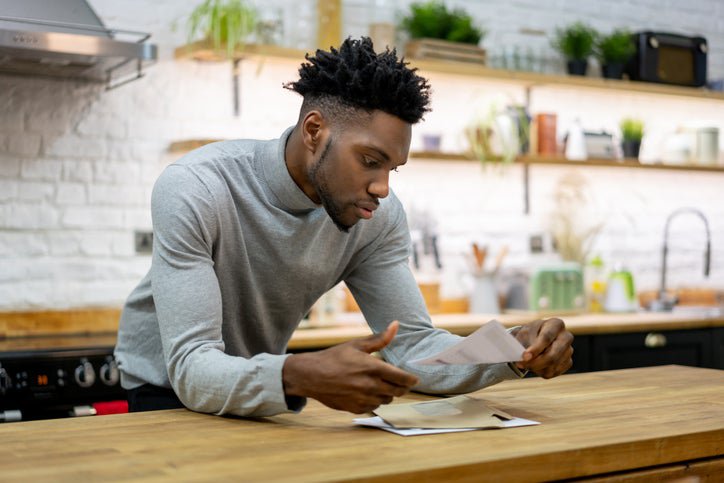 A man leaning against his kitchen counter while opening the mail.