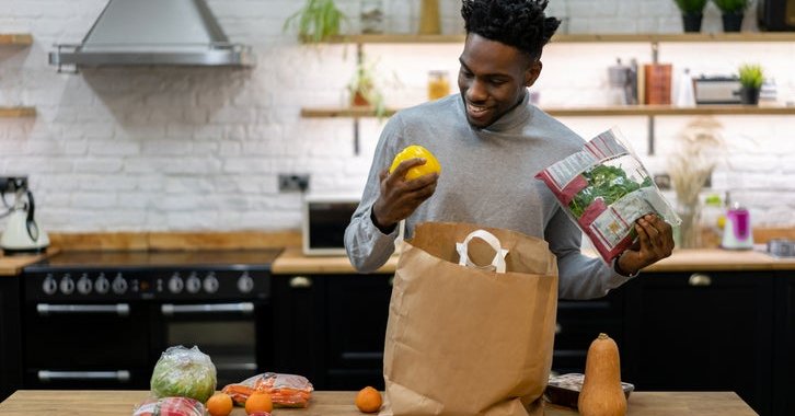 A smiling man standing in his kitchen and unpacking bags of groceries.