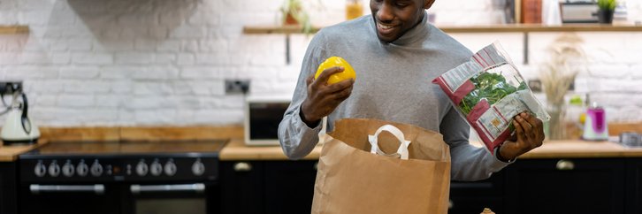 A smiling man standing in his kitchen and unpacking bags of groceries.