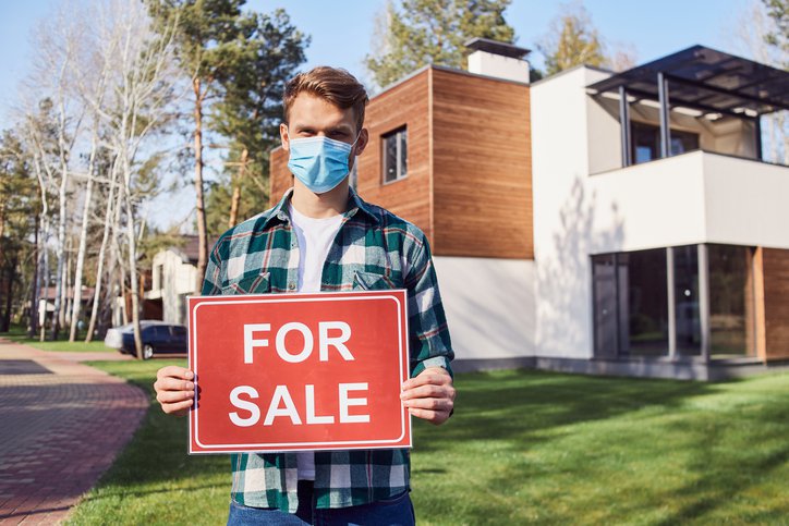 A man wearing a medical mask while standing in front of a home and holding a For Sale sign.