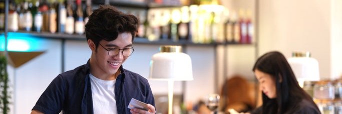 A man standing at a restaurant bar holding his phone and a credit card.