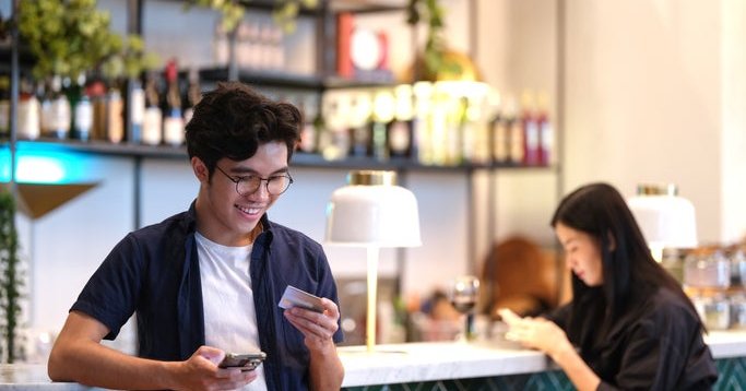 A man standing at a restaurant bar holding his phone and a credit card.