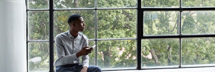 A man sitting next to a large window and holding a phone while looking thoughtfully outside.