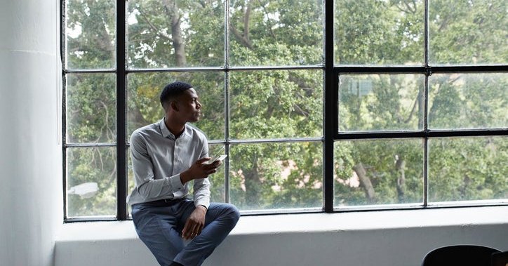 A man sitting next to a large window and holding a phone while looking thoughtfully outside.