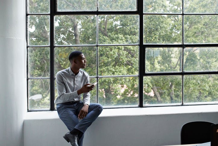 A man sitting next to a large window and holding a phone while looking thoughtfully outside.