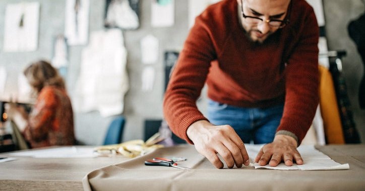 A man measuring and cutting fabric on a table with a woman working in the background.