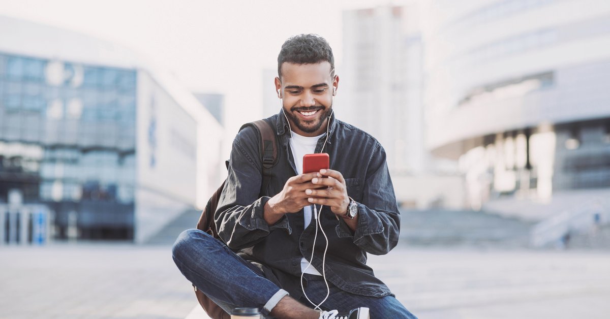 A man texting on his cell phone while sitting in an outdoor square between buildings.