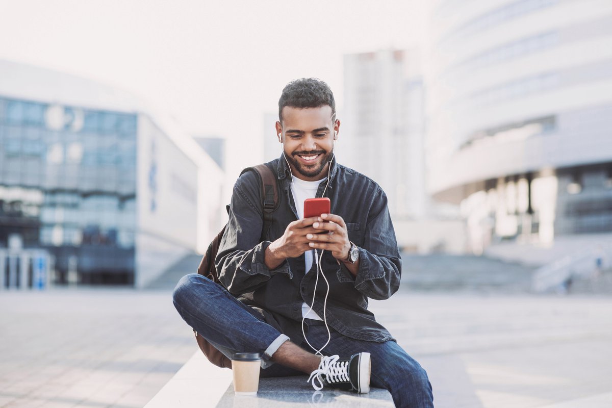 A man texting on his cell phone while sitting in an outdoor square between buildings.