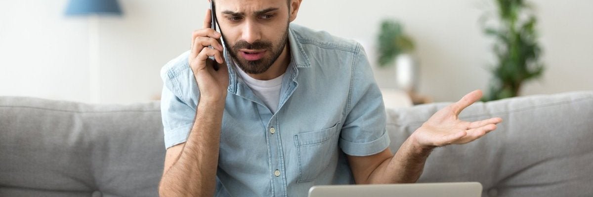 A man sitting on his couch and looking at his laptop in confusion while making a phone call.