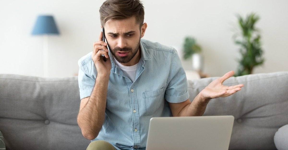 A man sitting on his couch and looking at his laptop in confusion while making a phone call.