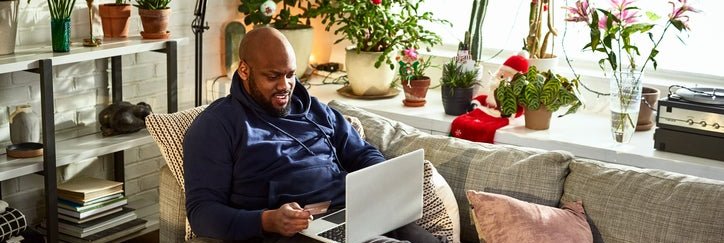 A man sitting on his couch surrounded by plants in a sunny room while online shopping on his laptop and holding a credit card in one hand.