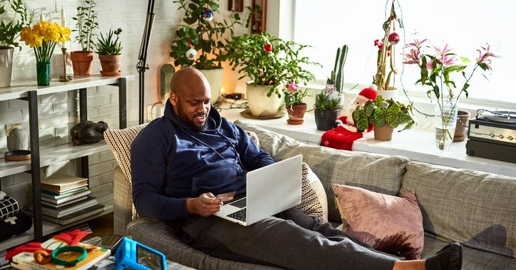 A man sitting on his couch surrounded by plants in a sunny room while online shopping on his laptop and holding a credit card in one hand.