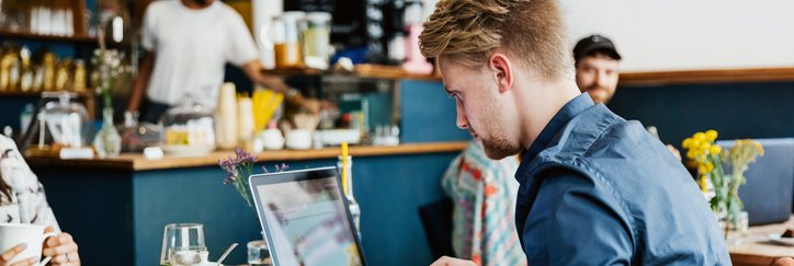 A man typing on a laptop while sitting at a table in a cafe.
