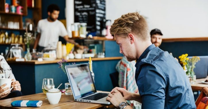 A man typing on a laptop while sitting at a table in a cafe.