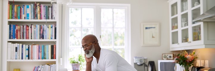 A man standing at the counter in his sunny kitchen and typing on his laptop.
