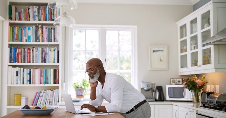 A man standing at the counter in his sunny kitchen and typing on his laptop.