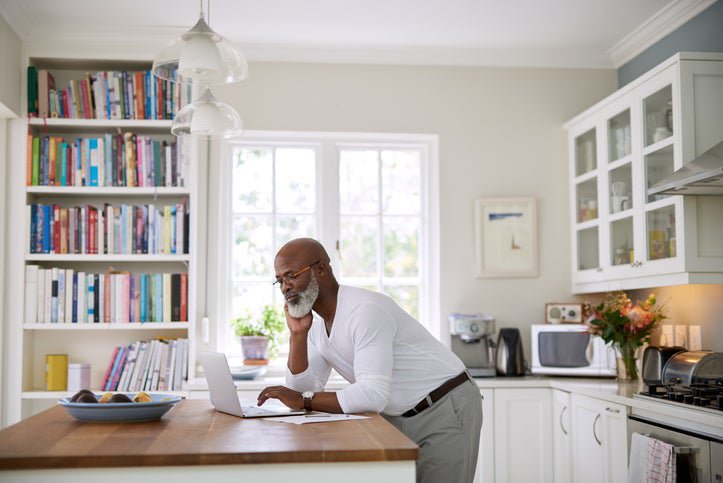 A man standing at his sunny kitchen counter and typing on his laptop.