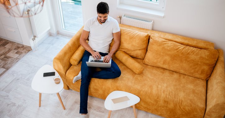 A man sitting on a couch in a sunny living room and typing on his laptop.