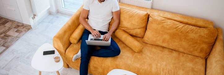 A man sitting on a couch in a sunny living room and typing on his laptop.
