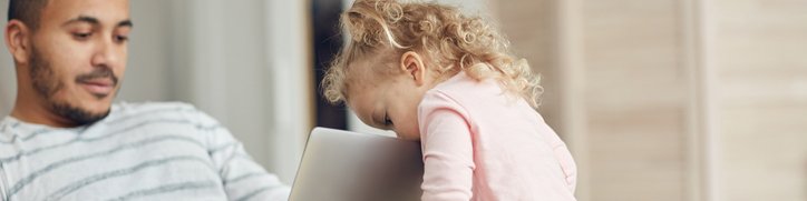 A man sitting on the couch and looking at his laptop while his toddler daughter peeks over the top of the screen.