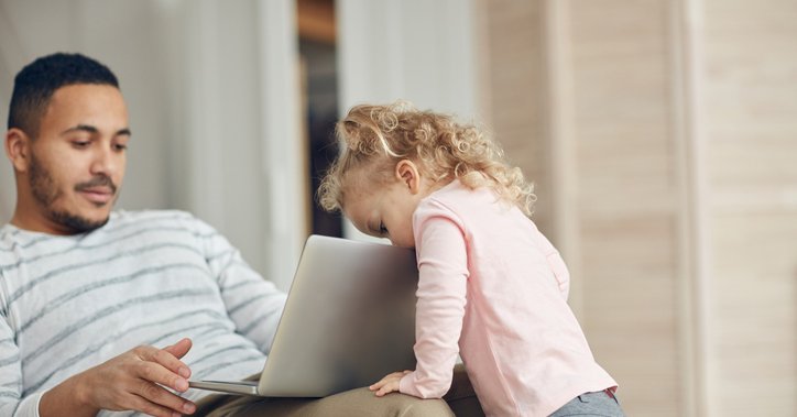 A man sitting on the couch and looking at his laptop while his toddler daughter peeks over the top of the screen.