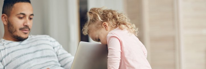 A man sitting on the couch and looking at his laptop while his toddler daughter peeks over the top of the screen.