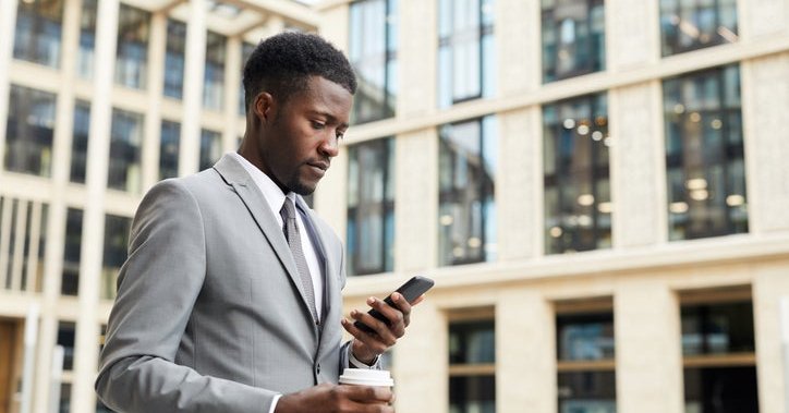 A man walking outside an office building while holding a coffee and looking at his phone.