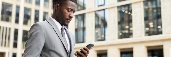 A man walking outside an office building while holding a coffee and looking at his phone.