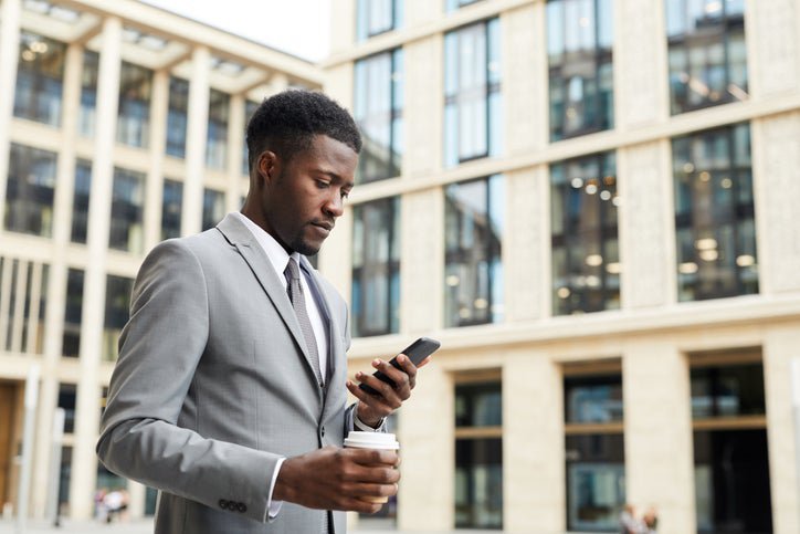 A man walking outside an office building while holding a coffee and looking at his phone.