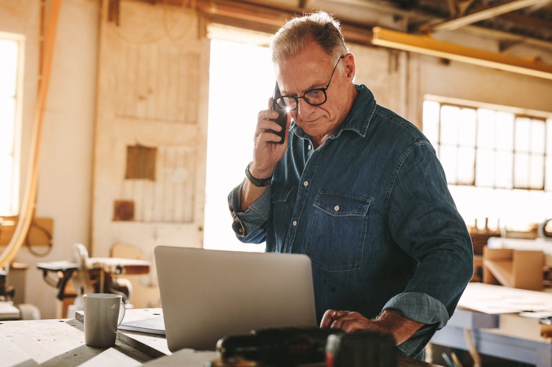 A man making a phone call while looking at his laptop in a sunny workshop.